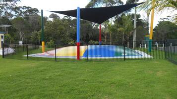 Children can play on the multi-level structure in this shaded outdoor play area surrounded by trees and green grass.