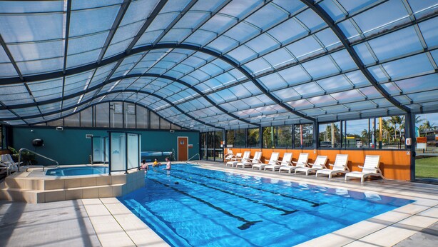 An indoor swimming pool with a glass ceiling, a changing room, and lounge chairs. - Tasman Holiday Parks – Merimbula (Merimbula)
