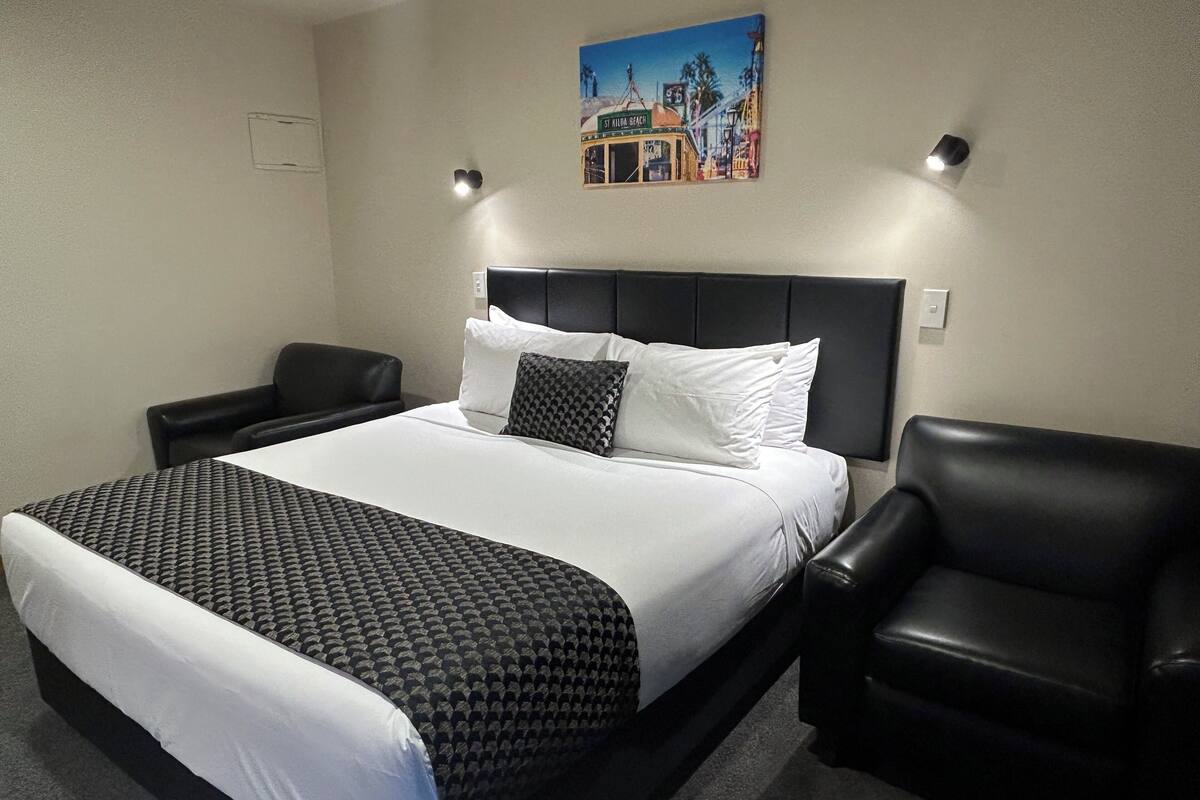 A hotel room with a large bed, two black leather chairs, a painting of a beach scene, and a nightstand.