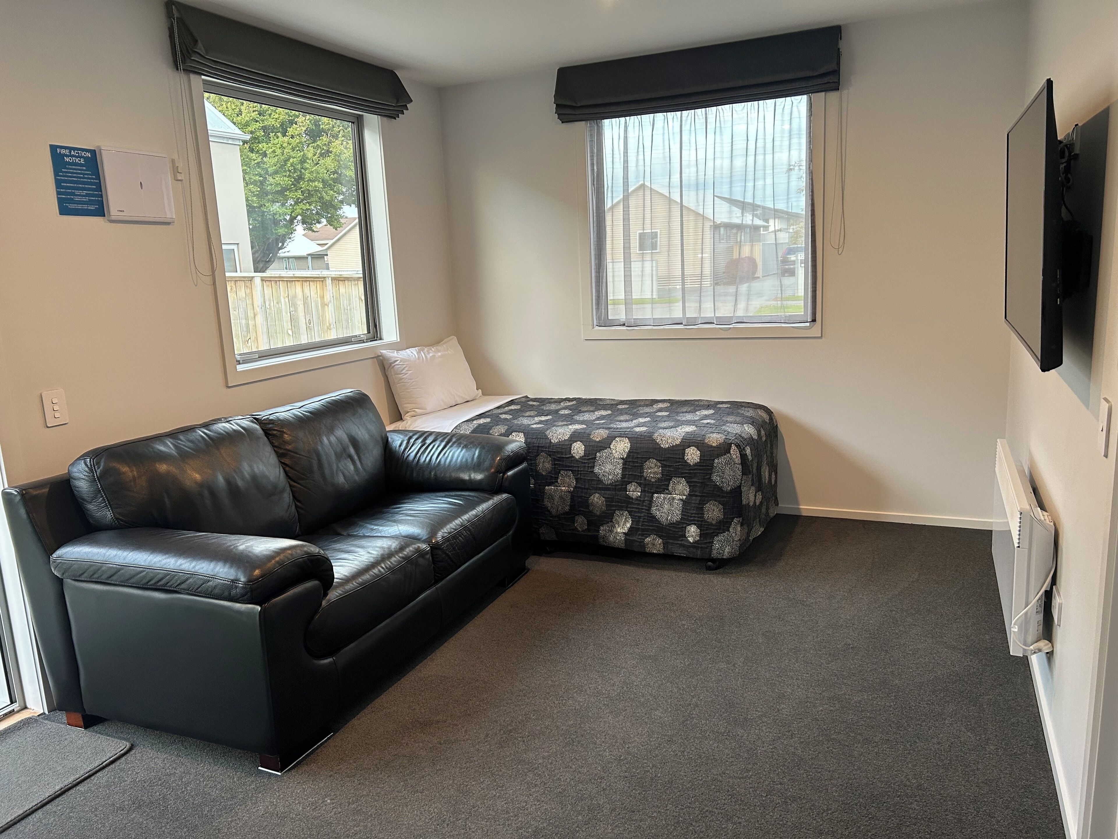 A small, furnished room with a black leather sofa, a bed with a patterned bedspread, two windows with blinds, a fire action notice on the wall, and a television mounted on the wall.
