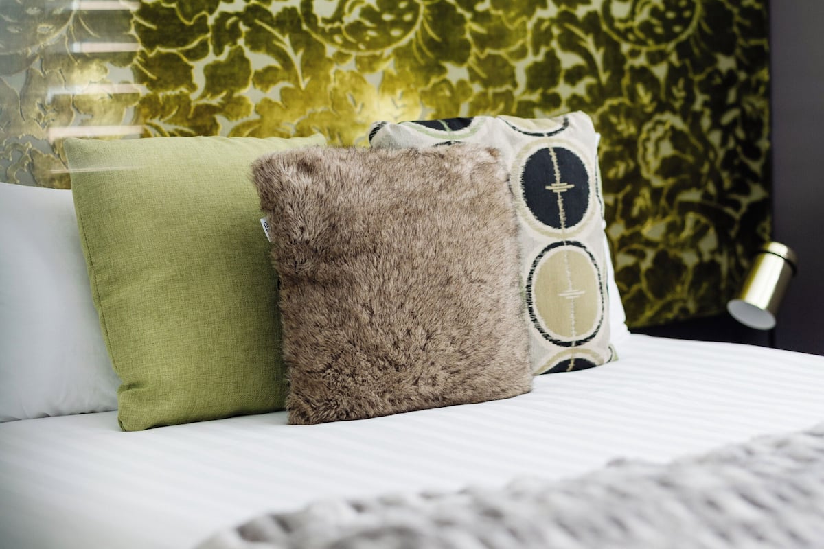A bed with a patterned headboard, a green pillow, a brown fluffy pillow, and a grey blanket.