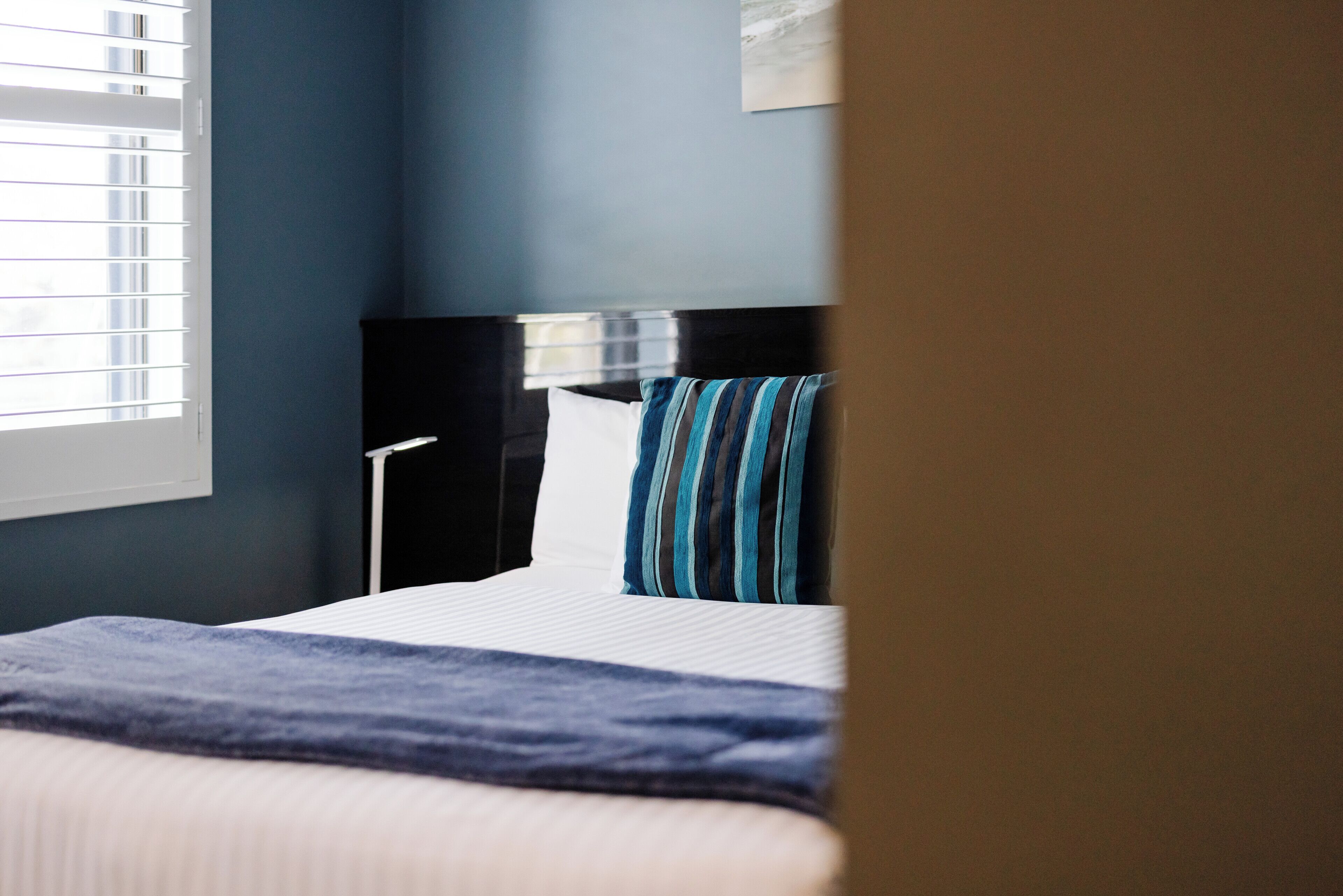 A hotel room with a bed, a nightstand, a lamp, and a window with blinds.