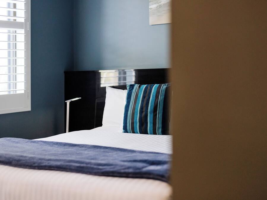 A hotel room with a bed, a nightstand, a lamp, and a window with blinds.