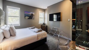 A bedroom with a bed, a TV, a glass shower enclosure, and a window with blinds.