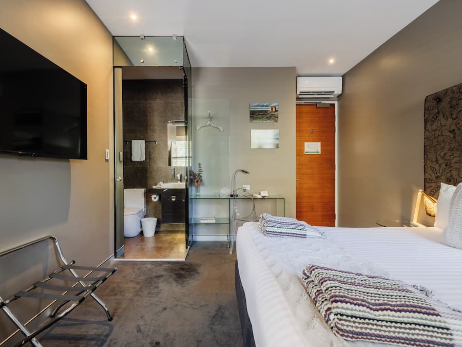 A modern hotel room with a large bed, a TV, a glass shower, and a wooden accent wall.