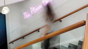 A staircase with a neon sign reading 'The Place to Stay' and a reflection of a smartphone on the glass railing.