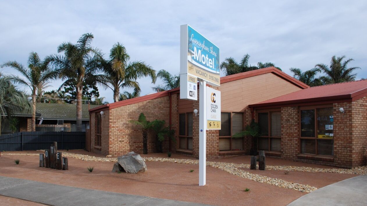 Um motel com fachada de tijolos, telhado vermelho e palmeiras ao fundo.