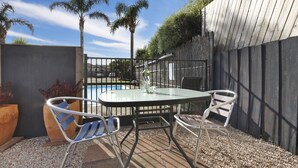 A patio with a round glass table and metal chairs, a pool in the background, and a wooden fence. - Cunningham Shore Motel (Lakes Entrance)