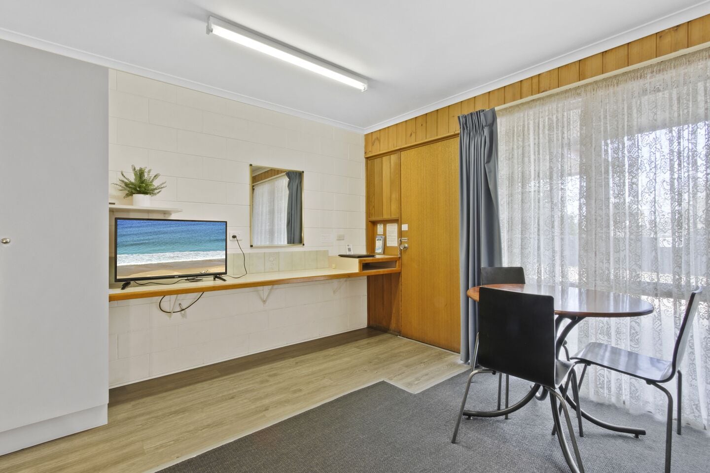 A compact room with a television on a shelf, a small table with chairs, a wooden door, and a window with sheer curtains.