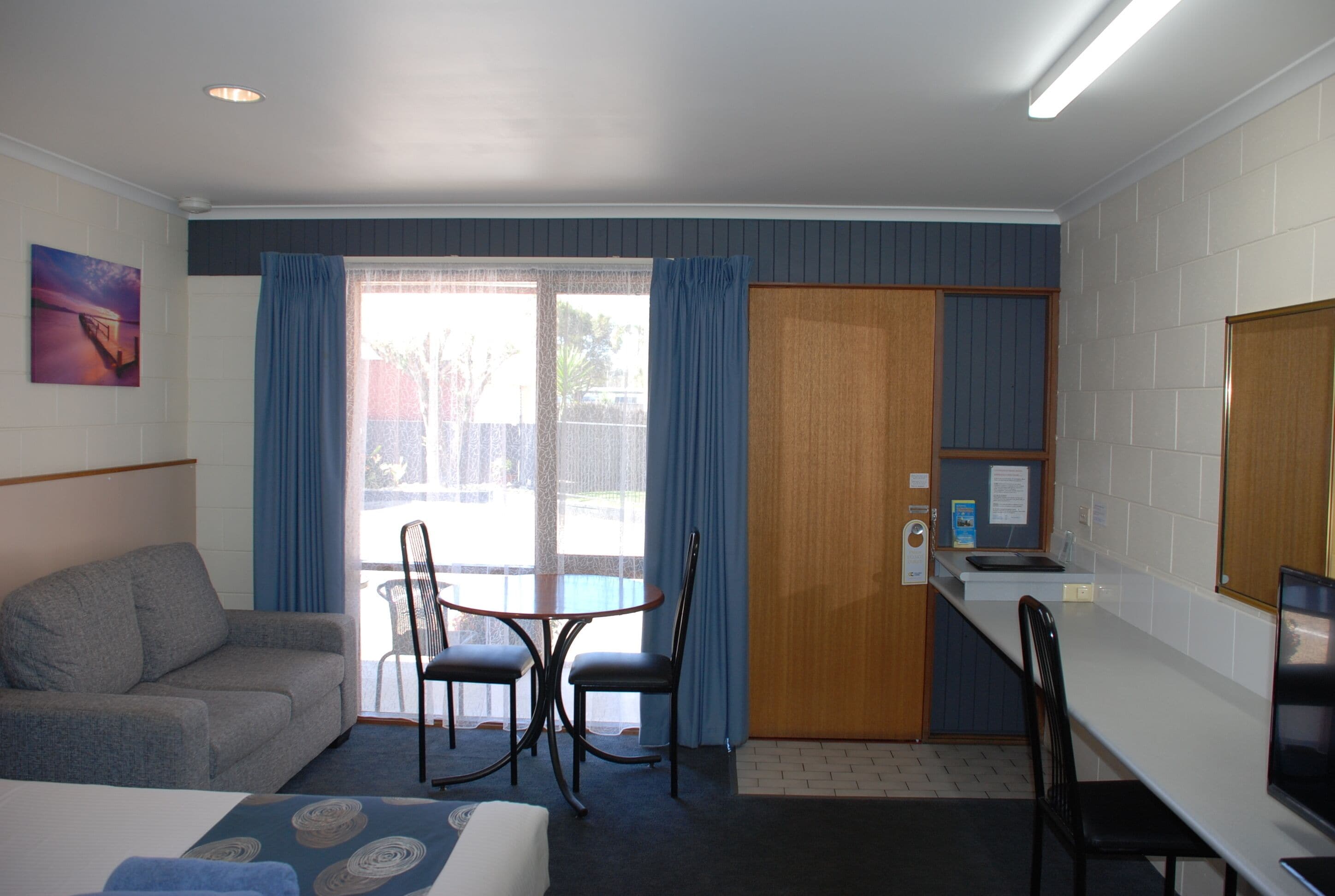 A hotel room with a sofa, a dining table with chairs, a kitchenette, and a sliding glass door to the outside.