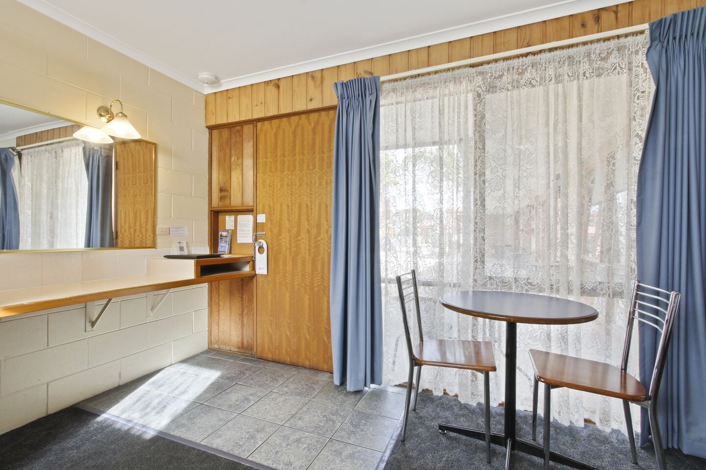 A compact kitchen with a wooden countertop, a sink, and a mirror. There is a round table with two chairs and a window with lace curtains.