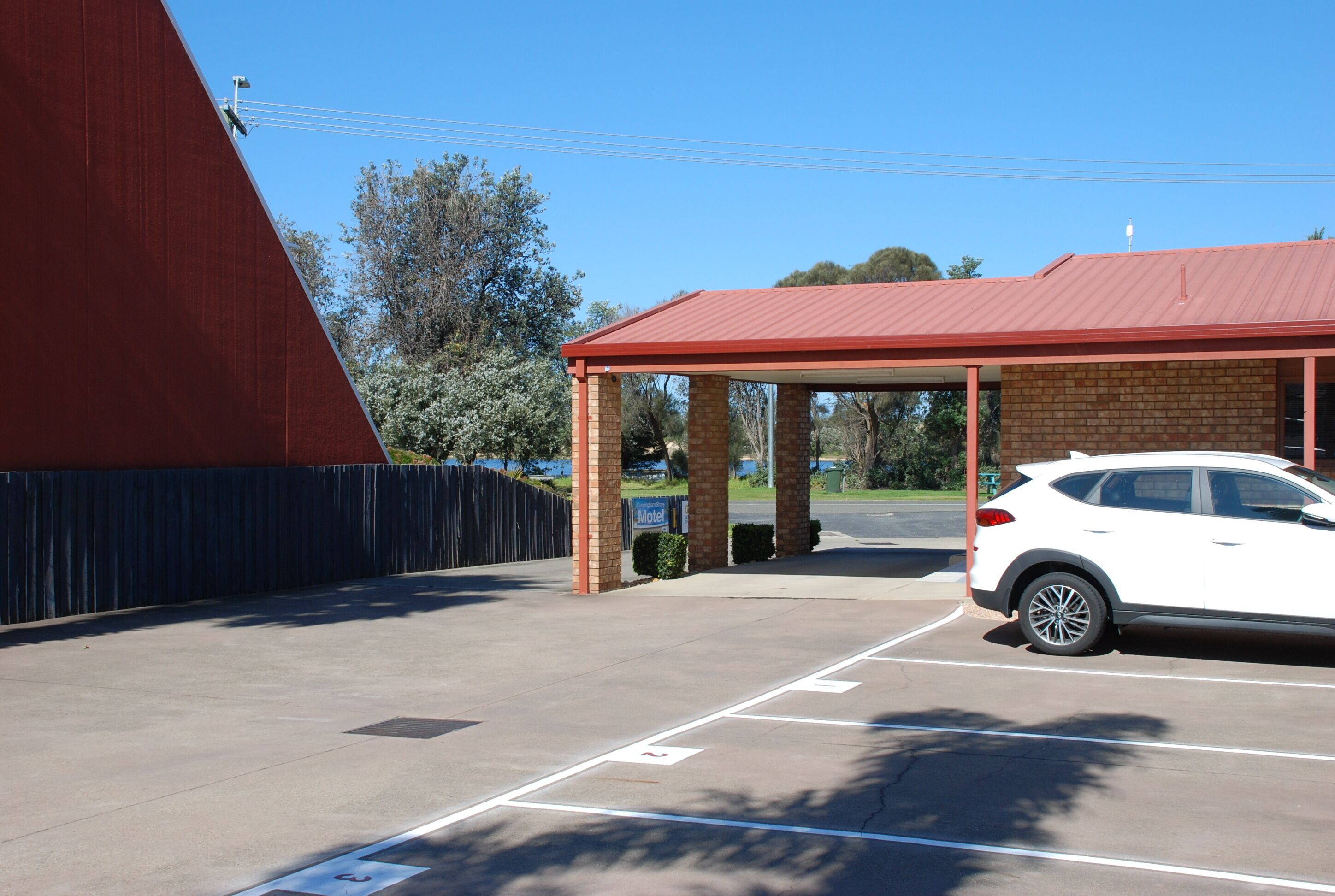 Un estacionamiento con un automóvil blanco aparcado en un espacio asignado, una zona cubierta con techo rojo y un edificio de ladrillo al fondo.