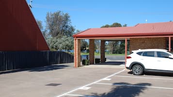 Un estacionamiento con un automóvil blanco aparcado en un espacio asignado, una zona cubierta con techo rojo y un edificio de ladrillo al fondo.