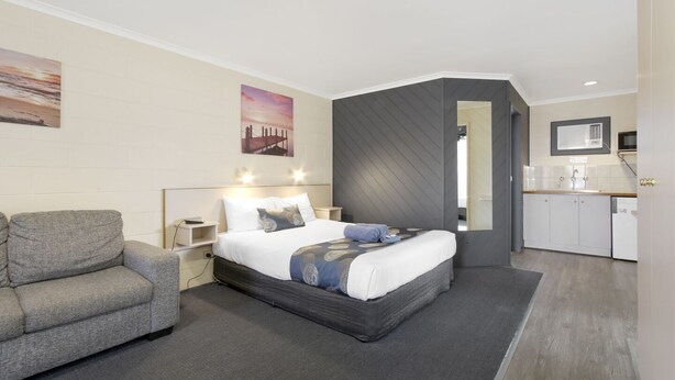 A modern hotel room with a bed, a grey sofa, a small bedside table, a nightstand, a wall-mounted TV, and a kitchenette with a microwave and sink. - Cunningham Shore Motel (Lakes Entrance)