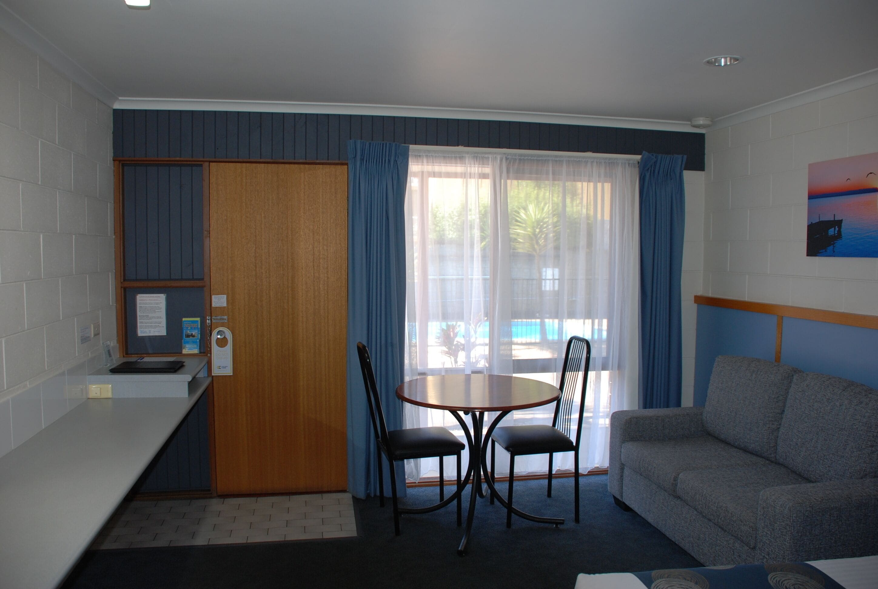 A compact hotel room with a kitchenette, a dining area with a table and chairs, a sofa, and a large window with curtains.