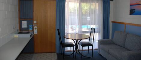 A compact hotel room with a kitchenette, a dining area with a table and chairs, a sofa, and a large window with curtains.