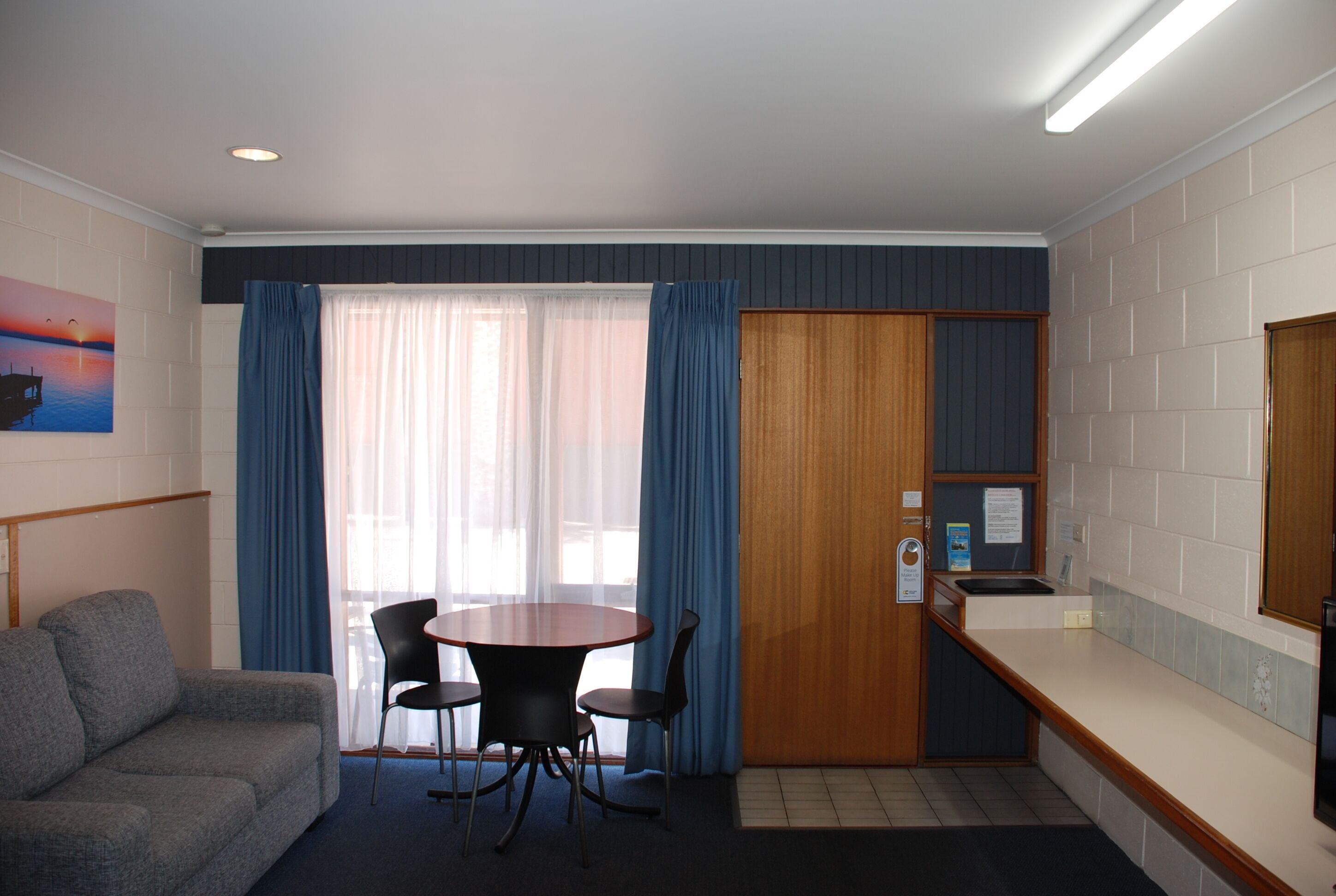 A compact hotel room with a sofa, a round table with chairs, a wooden door, and a small kitchenette.