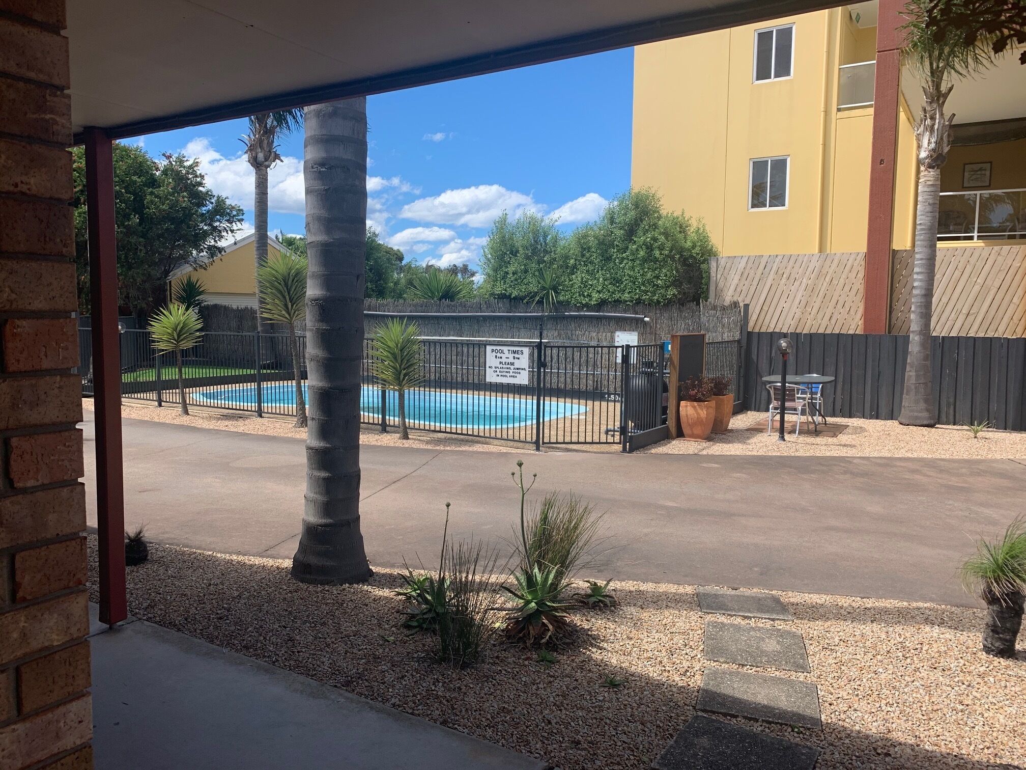 Vue d’une piscine entourée d’une clôture, avec des palmiers et un bâtiment en arrière-plan.