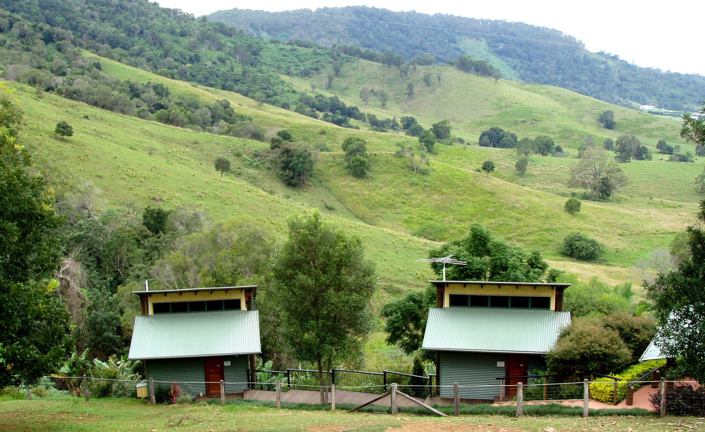 Two small, single-story buildings with green roofs, surrounded by a fence, set against a backdrop of rolling hills and scattered trees.