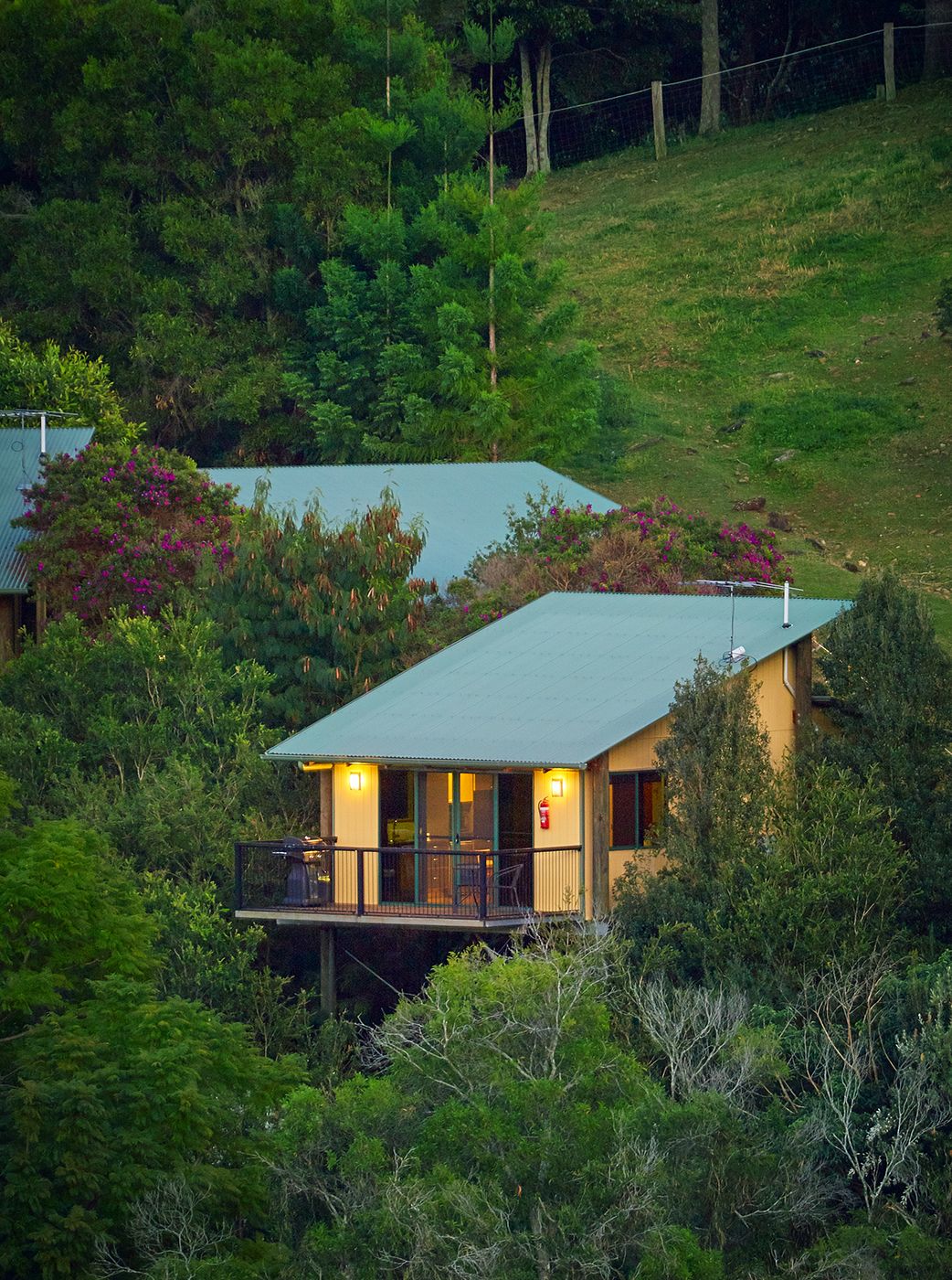 Une maison à deux étages dotée d’un balcon, entourée d’une végétation luxuriante et d’arbres.