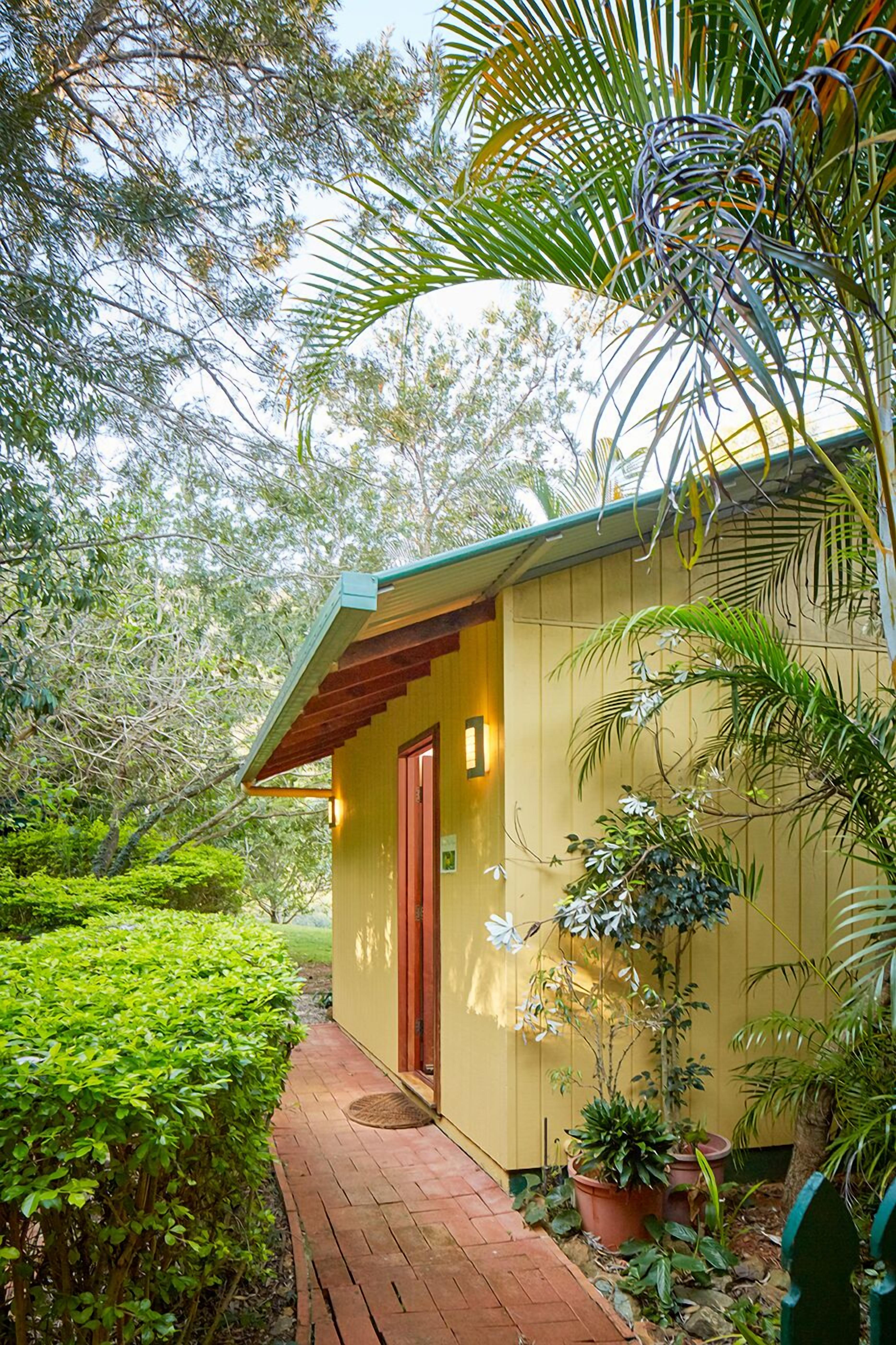 Un petit bâtiment d’un seul étage, avec une porte rouge et un toit vert, entouré d’une végétation luxuriante et de plantes en pot.