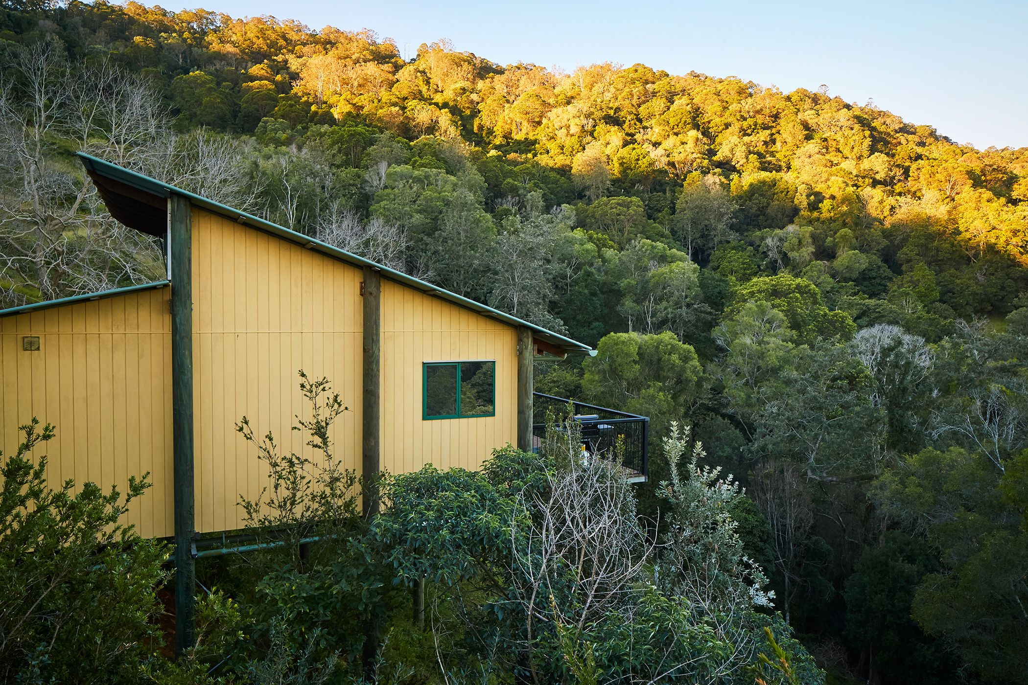A yellow building with a green roof, surrounded by dense forest and hills.