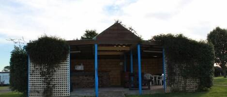 A small wooden structure with a pitched roof, possibly a gazebo or a small shelter, surrounded by a white lattice fence and some greenery.