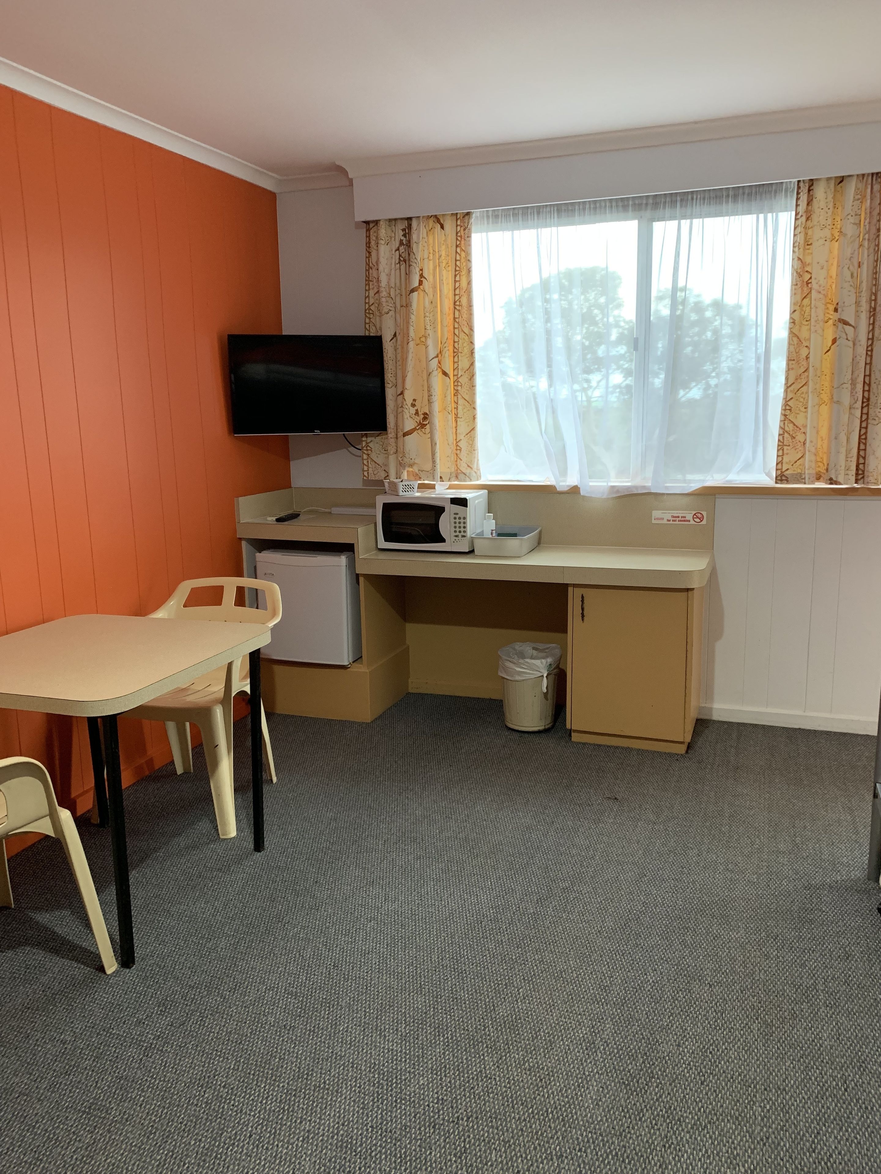 Een kleine kamer met een aan de muur bevestigde televisie, een magnetron op een klein tafeltje, een bureau met stoel, en een raam met gordijnen.