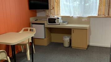 Een kleine kamer met een aan de muur bevestigde televisie, een magnetron op een klein tafeltje, een bureau met stoel, en een raam met gordijnen.
