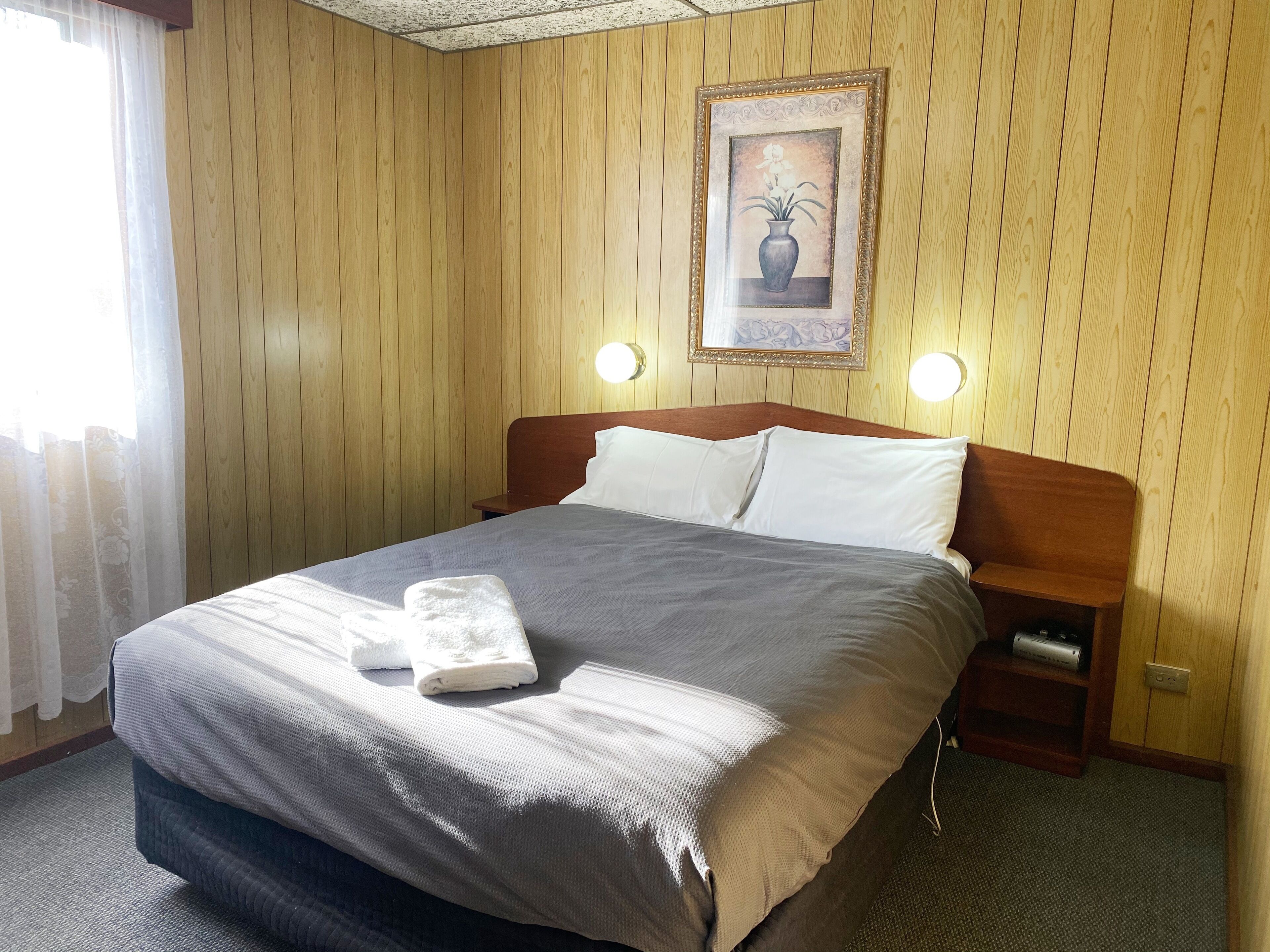 A hotel room with a wooden paneled wall, a bed with a grey bedspread and white pillows, a framed picture on the wall, and two bedside lamps.