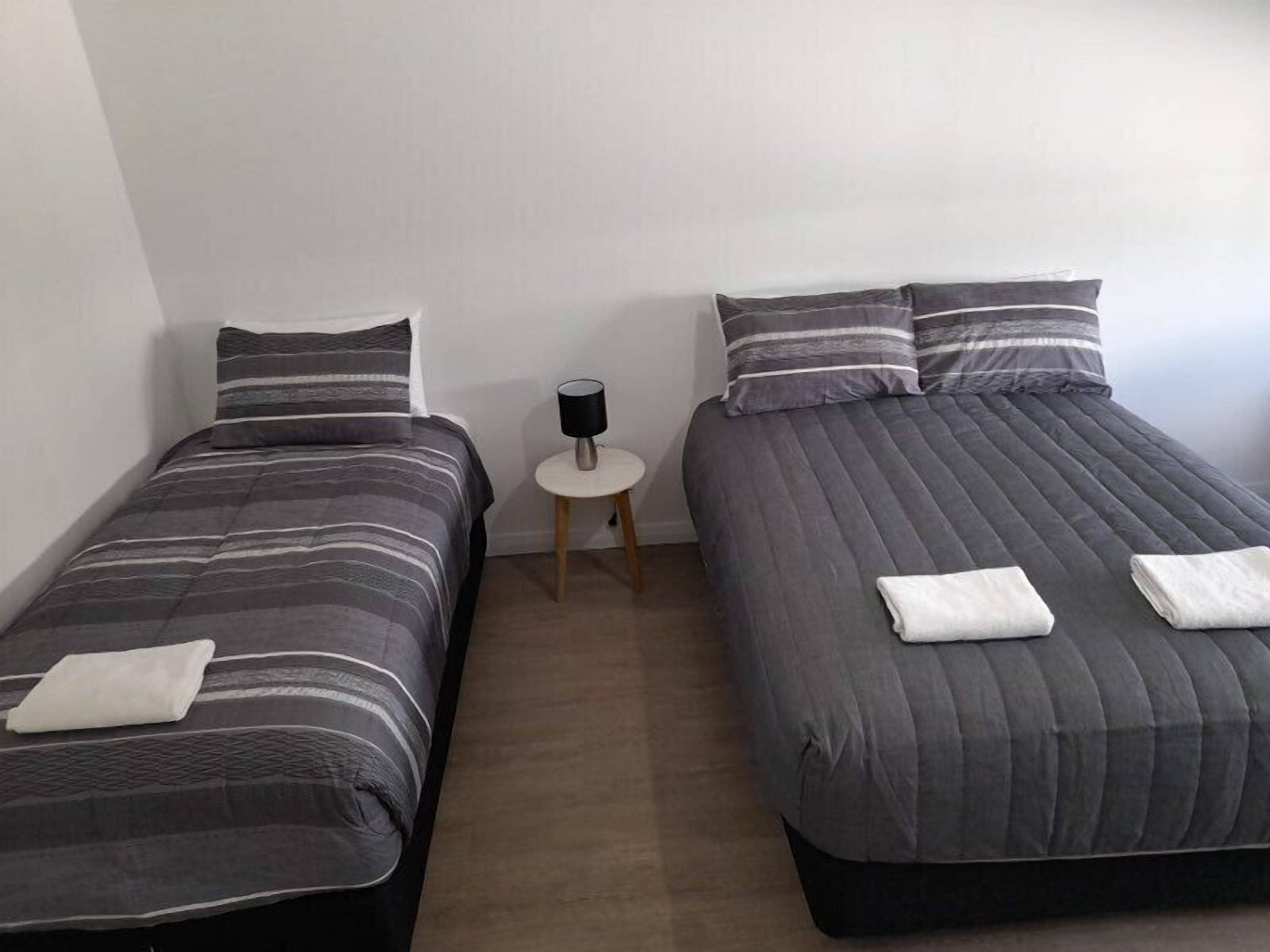Zwei Einzelbetten mit grauer Bettwäsche und gestreiften Kissen, ein kleiner runder Tisch mit einer Lampe und eine weiße Wand.