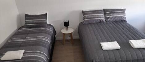 Zwei Einzelbetten mit grauer Bettwäsche und gestreiften Kissen, ein kleiner runder Tisch mit einer Lampe und eine weiße Wand.