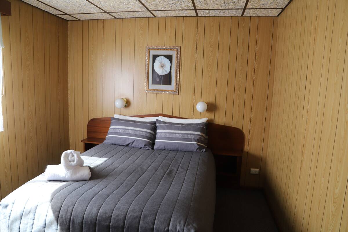Een hotelkamer met een houten lambrisering, een bed met grijze beddengoed, een ingelijst schilderij aan de muur en twee bedlampjes.