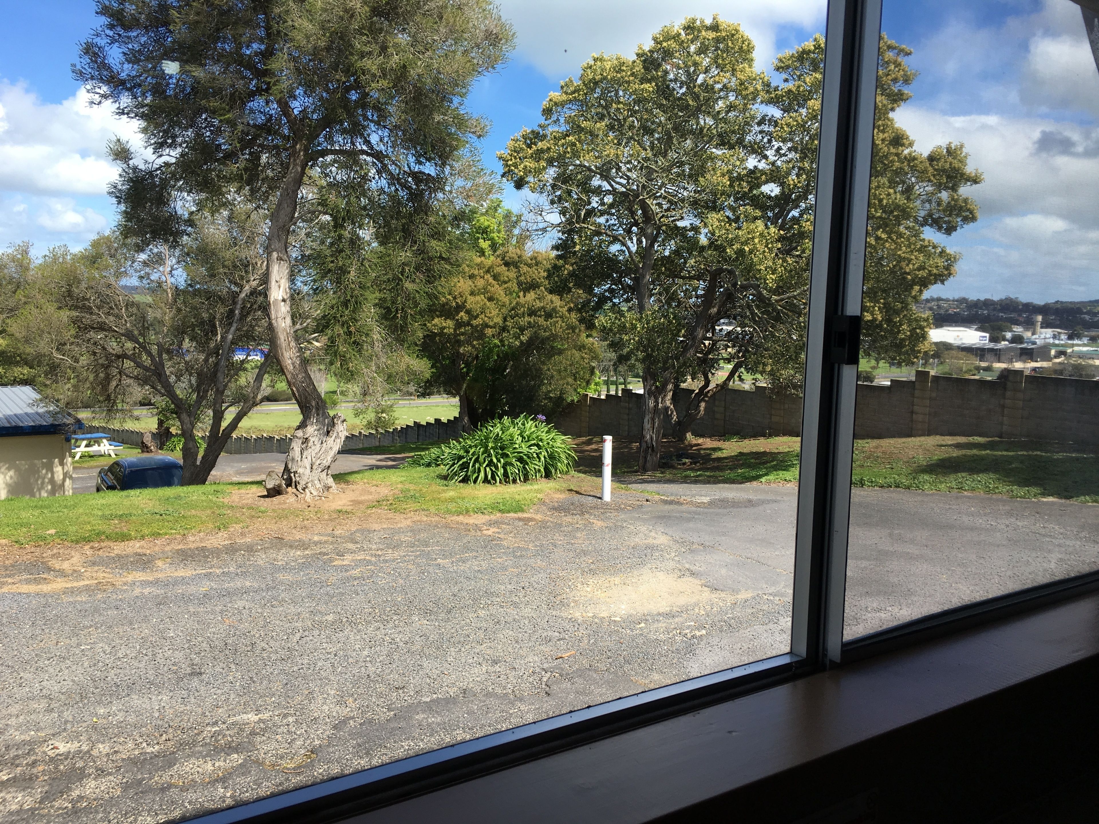 A view from a window of a parking area with trees, a picnic table, and a gravel path.