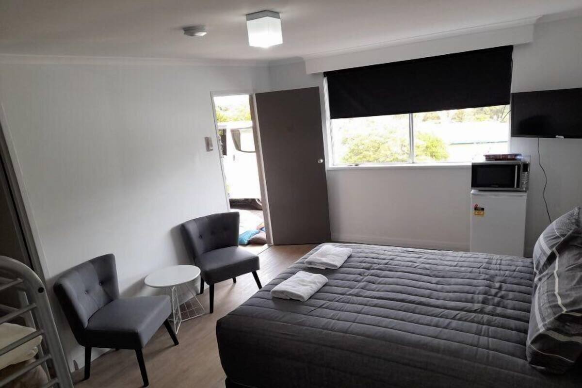 Een slaapkamer met een bed, twee stoelen, een klein tafeltje, een raam met een donkerblinde en een magnetron op een kast.