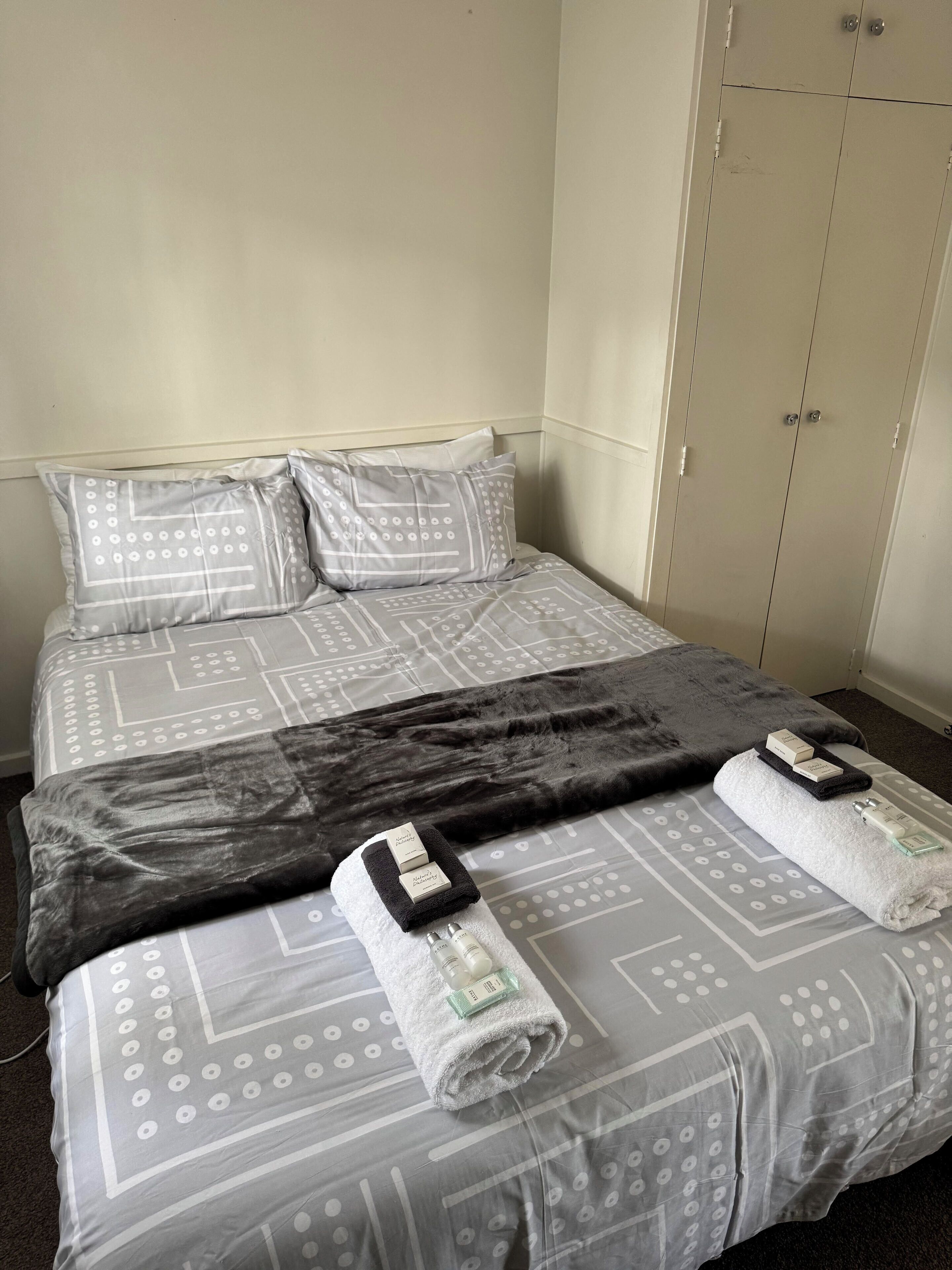 Una cama bien tendida con ropa de cama estampada, un plaid oscuro y dos toallas con artículos de aseo.