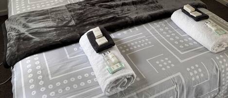 Una cama bien tendida con ropa de cama estampada, un plaid oscuro y dos toallas con artículos de aseo.