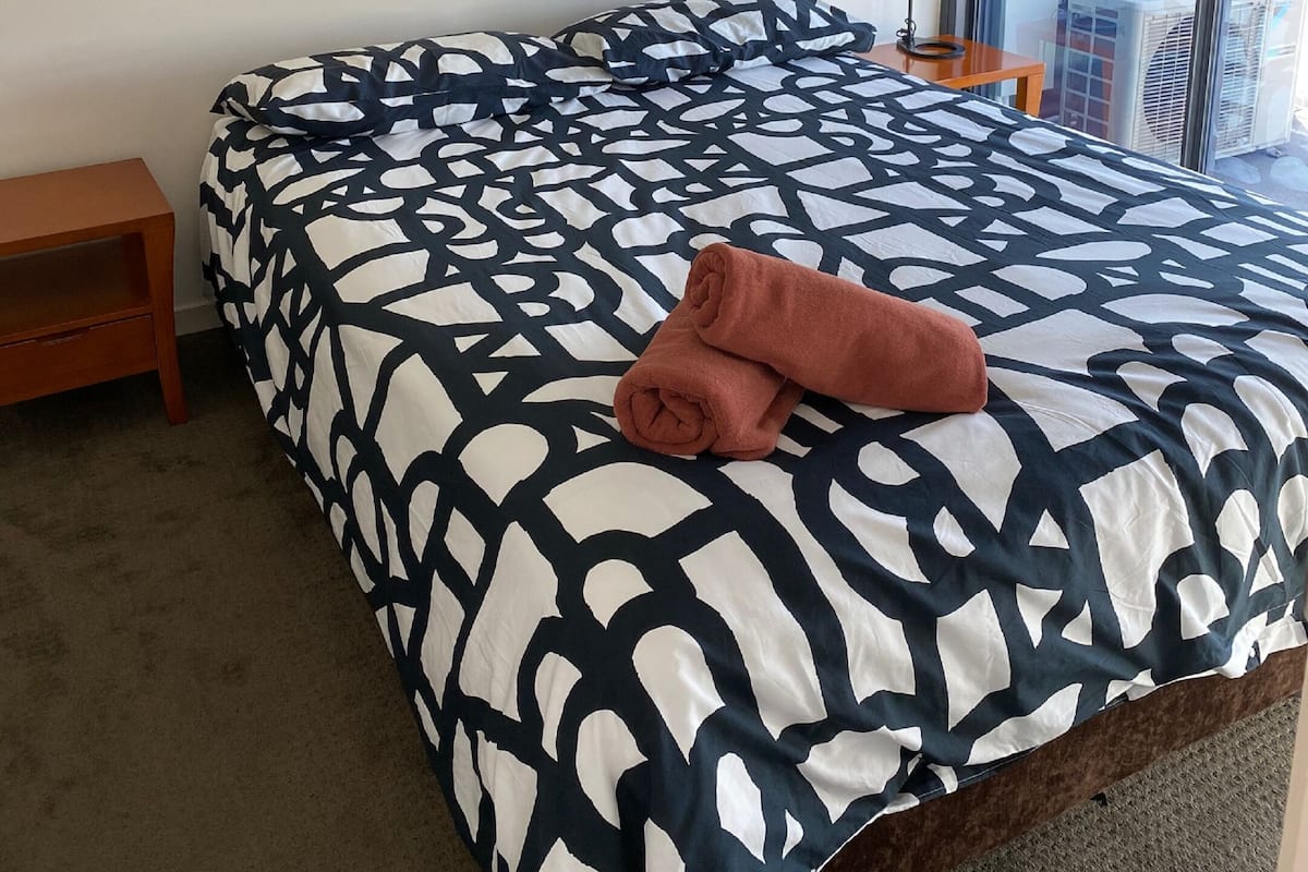 Ein ordentlich bezogenes Bett mit einem geometrisch gemusterten Überwurf, ein kleiner Nachttisch aus Holz, ein gerahmtes Kunstwerk an der Wand und ein Fenster mit Blick auf Gebäude.