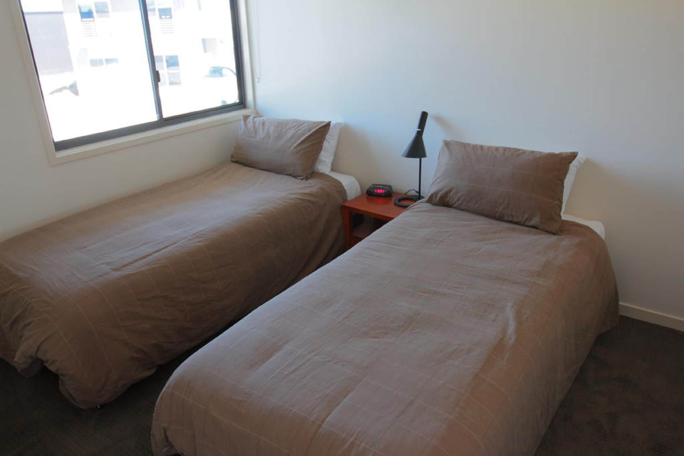A room with two single beds, a nightstand with a lamp, and a window providing a view of buildings outside.
