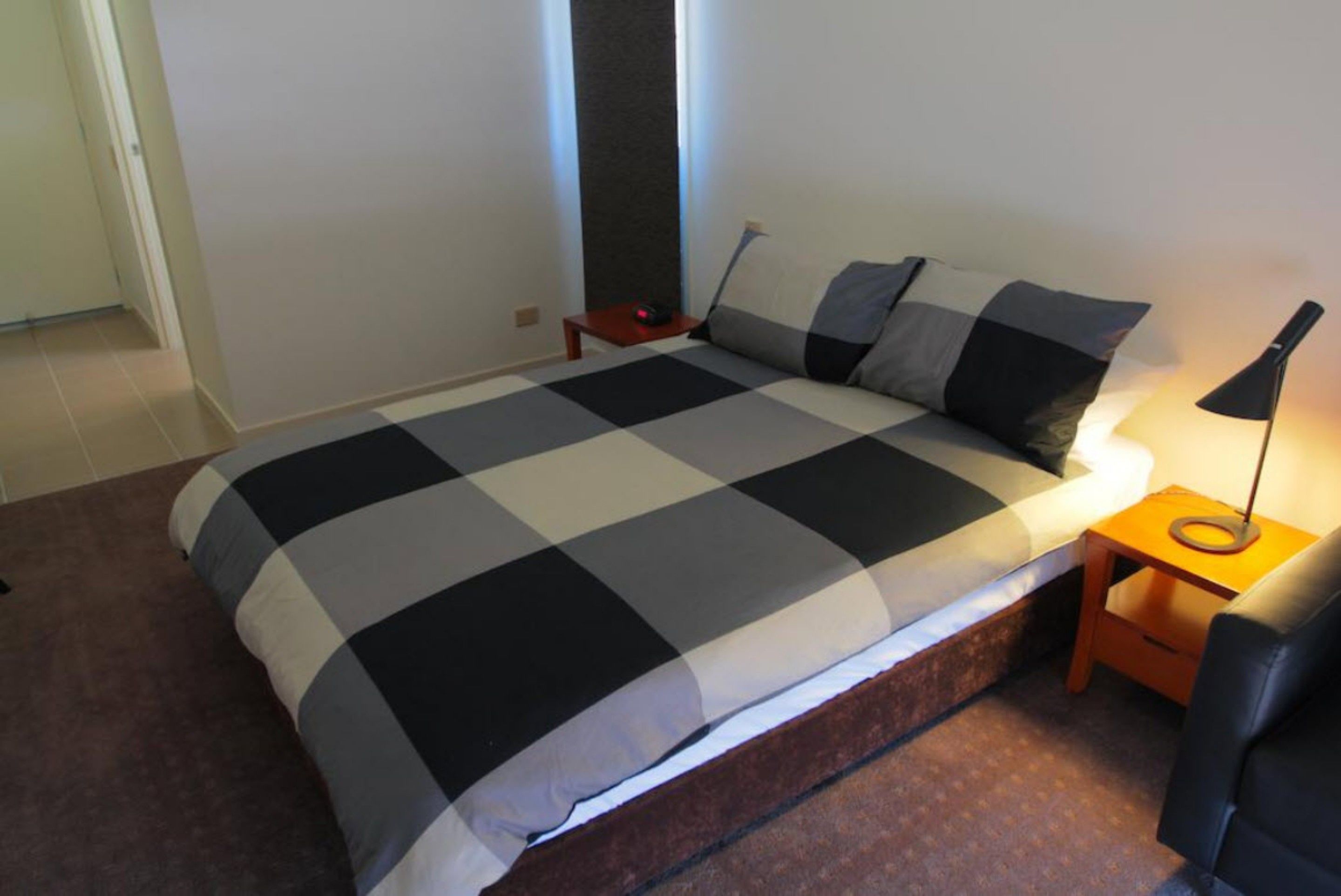 Un dormitorio con una colcha a cuadros, una mesita de noche con lámpara y una puerta al fondo.