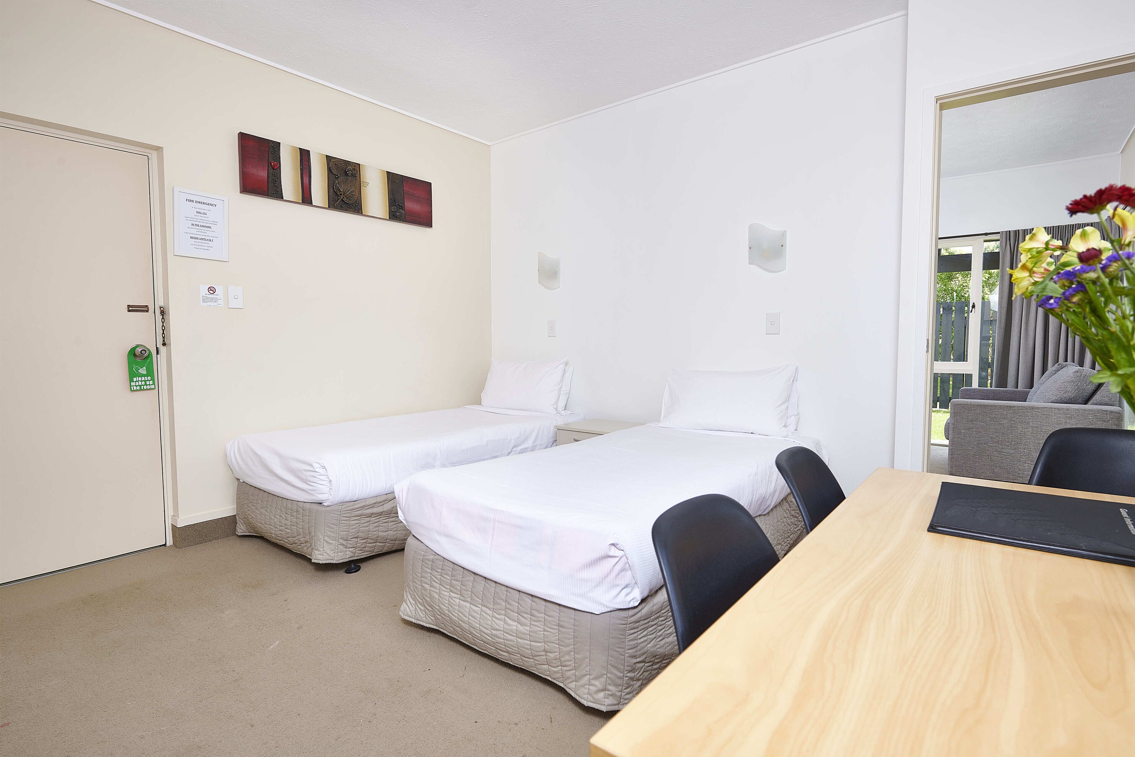 A room with two single beds, a wooden table, and a chair. There is a door with a fire emergency exit sign, and a small wall decoration above the beds.