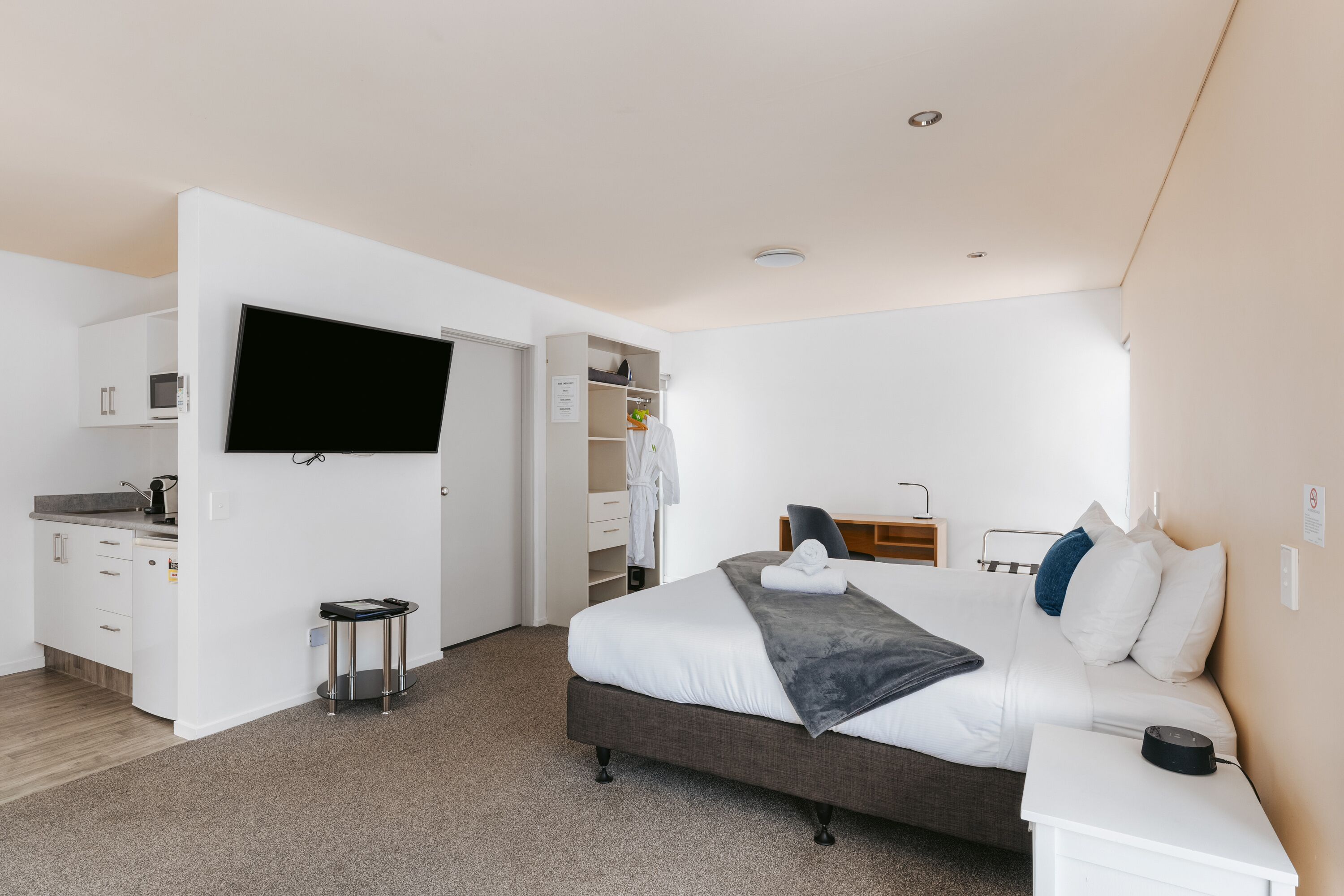 A modern hotel room with a large bed, a flat-screen TV mounted on the wall, a small kitchenette, and a walk-in closet.