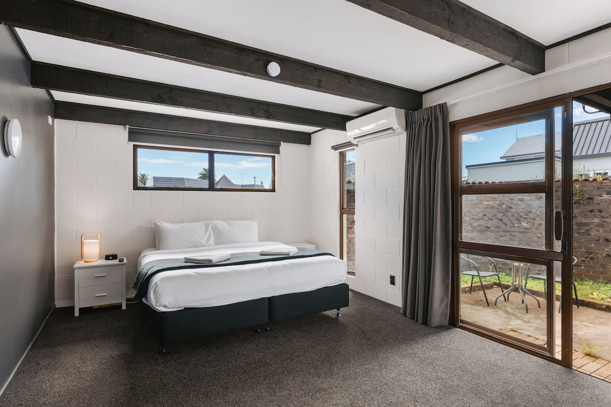 A modern bedroom with a large bed, a bedside table with a lamp, a window, and a sliding glass door leading to an outdoor area.