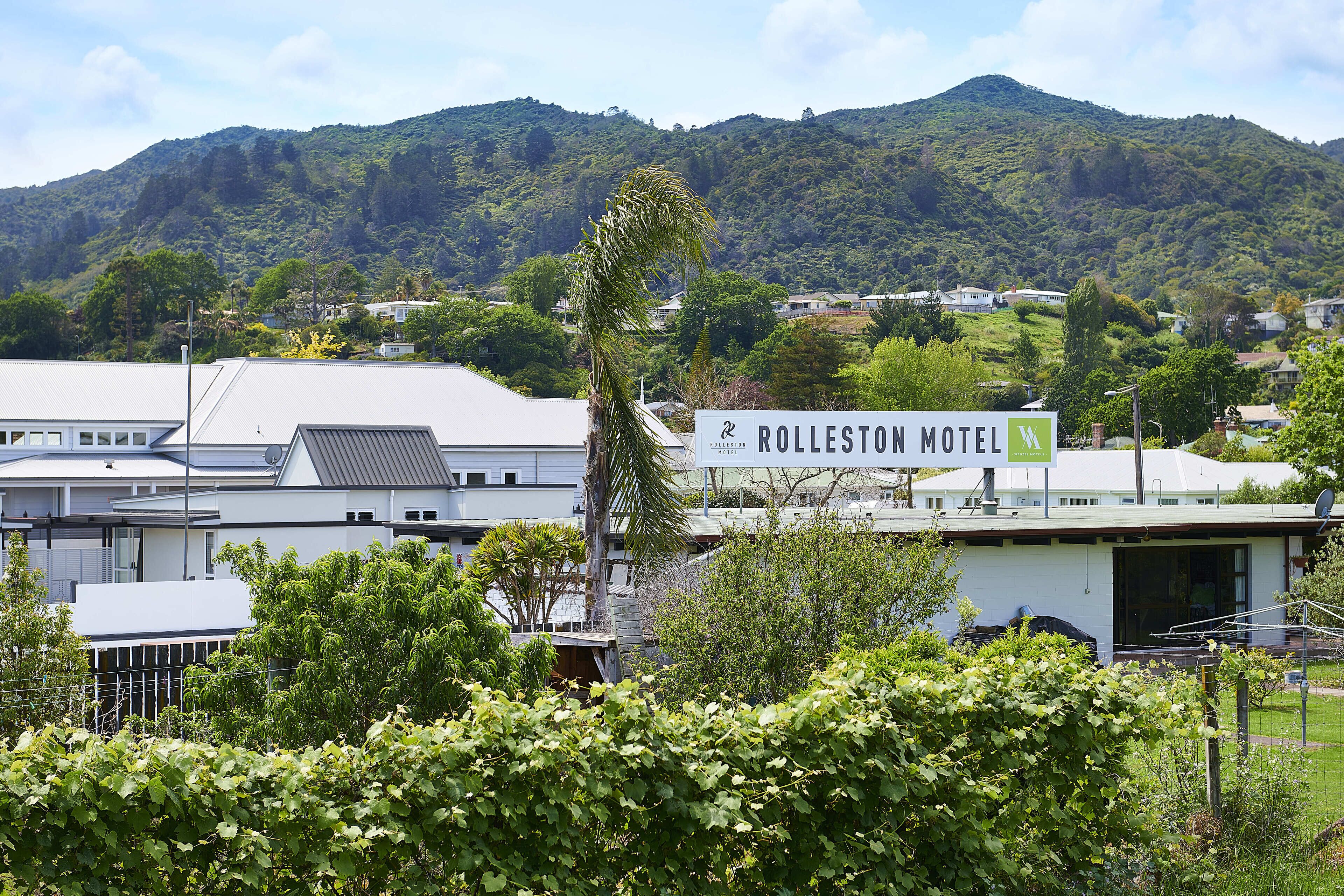 A hotel with a sign reading "Rolleston Motel" amidst lush greenery and hills in the background.