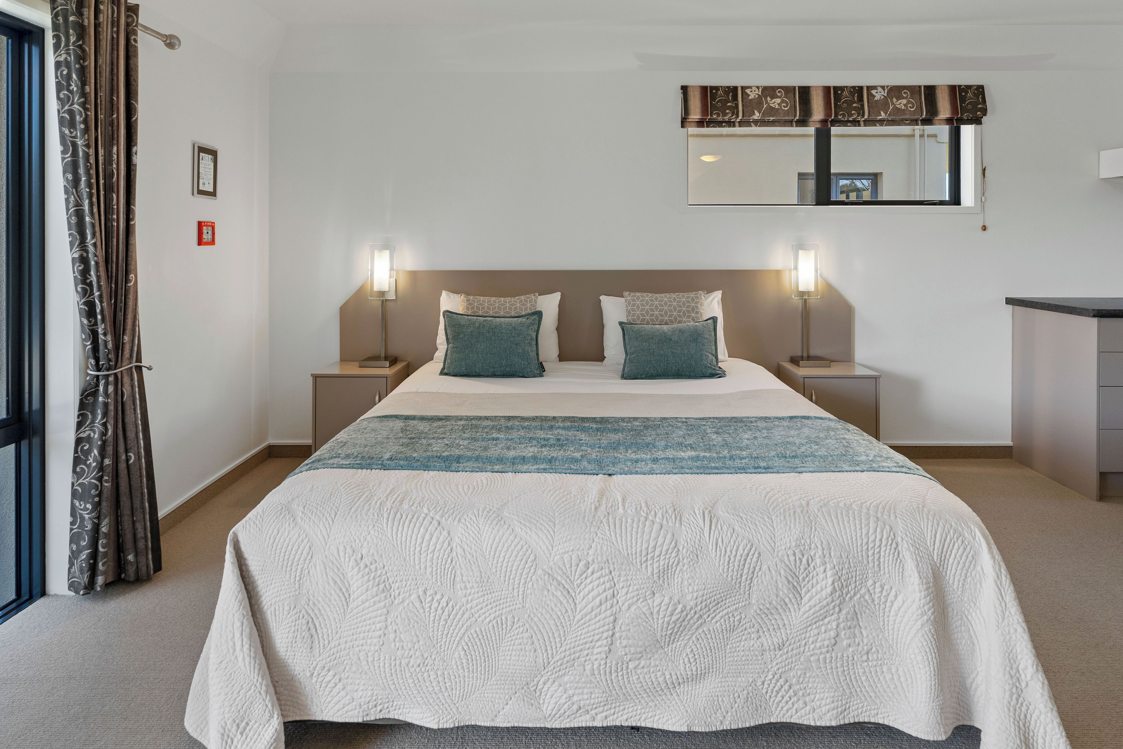A hotel room with a large bed, two bedside tables, a nightstand, a window with curtains, and a small desk.