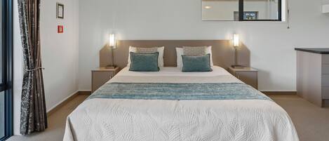 Habitación de hotel con una cama grande, dos mesitas de noche, un buró, una ventana con cortinas y un escritorio pequeño.