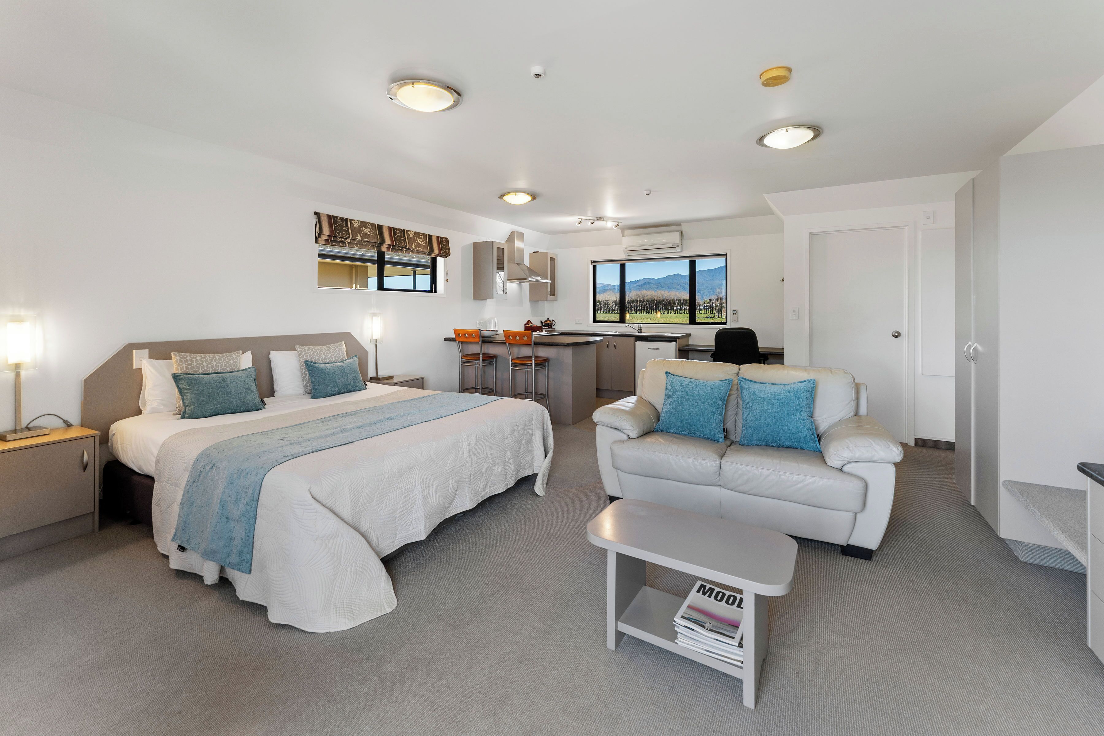 A modern hotel room with a bed, a sofa, a small table, and a kitchenette.