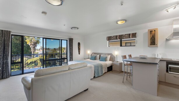 A modern hotel room with a balcony, a sofa, a bed, a kitchen area, and a dining table. - Avalon Manor Motel (Motueka)