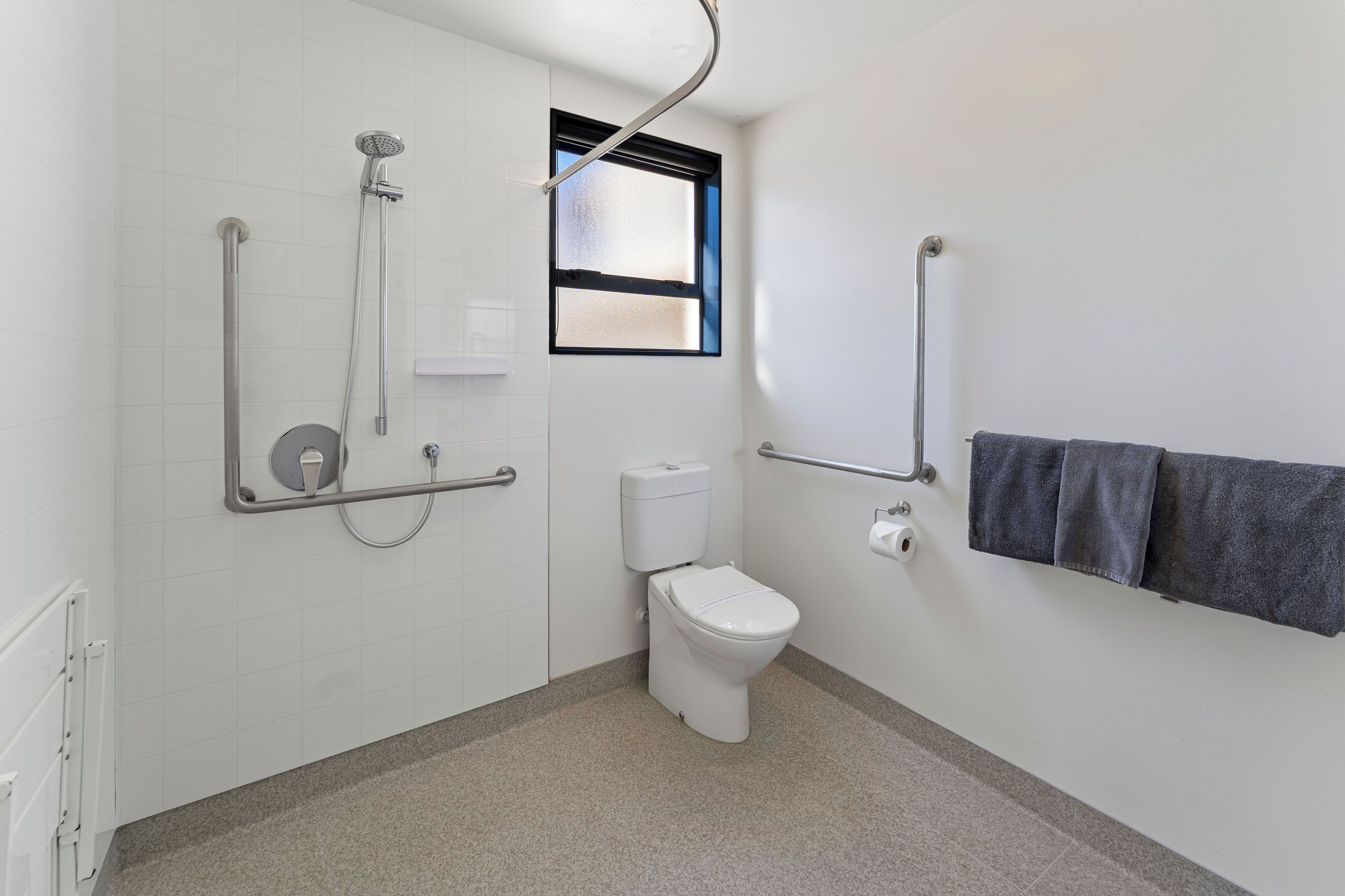 Un baño con inodoro, barras de apoyo, ducha y ventana.