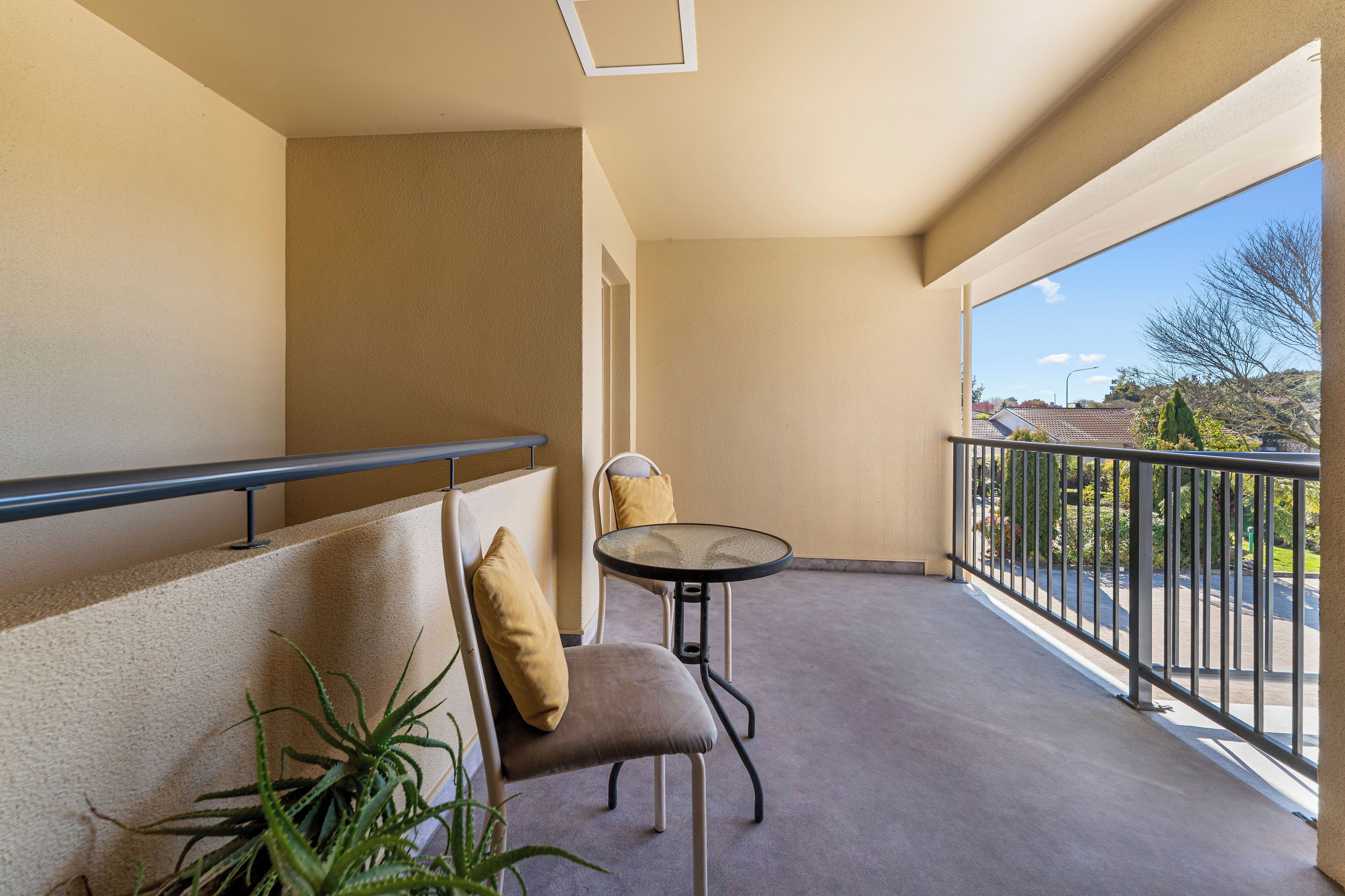 A balcony with a chair and table, beige walls, and a view of a parking lot and buildings.